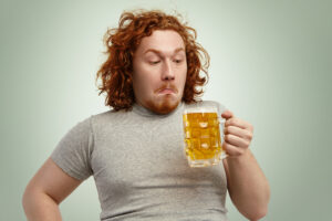 Clueless red haired young man with curly hair holding glass of light beer, looking at it, having confused indecisive expression, hesitating, thinking drink it or not, standing against blank wall