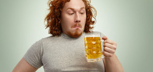 Clueless red haired young man with curly hair holding glass of light beer, looking at it, having confused indecisive expression, hesitating, thinking drink it or not, standing against blank wall